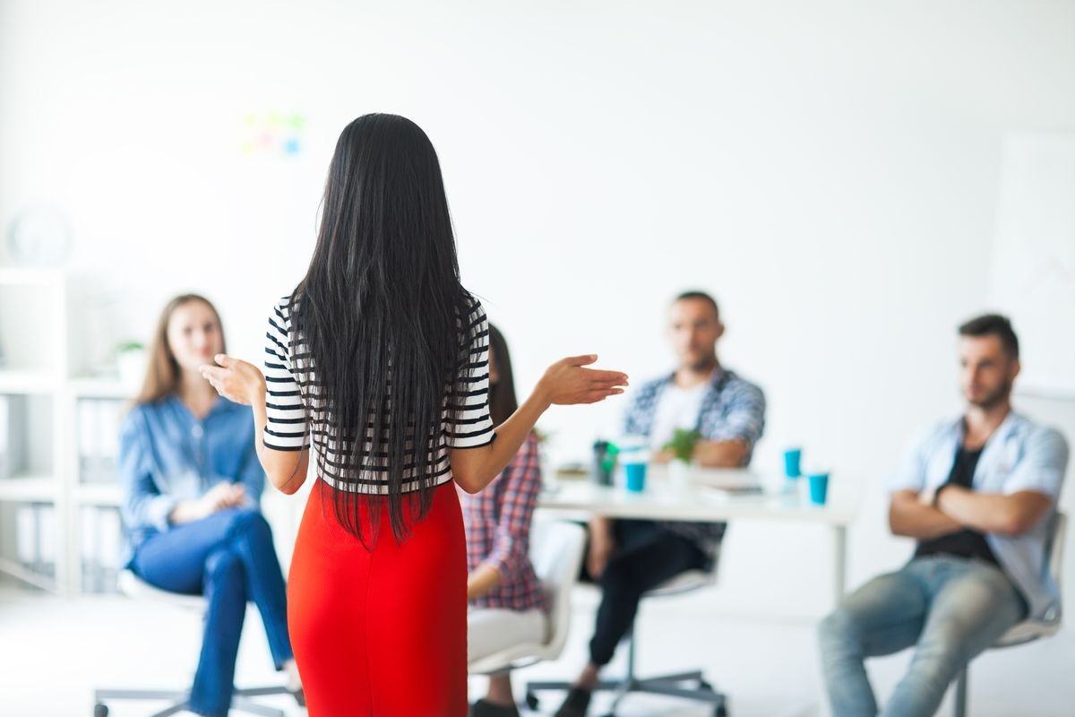  woman business coach gesturing with hand while standing against defocused group of people sitting at the chairs in front of her leader